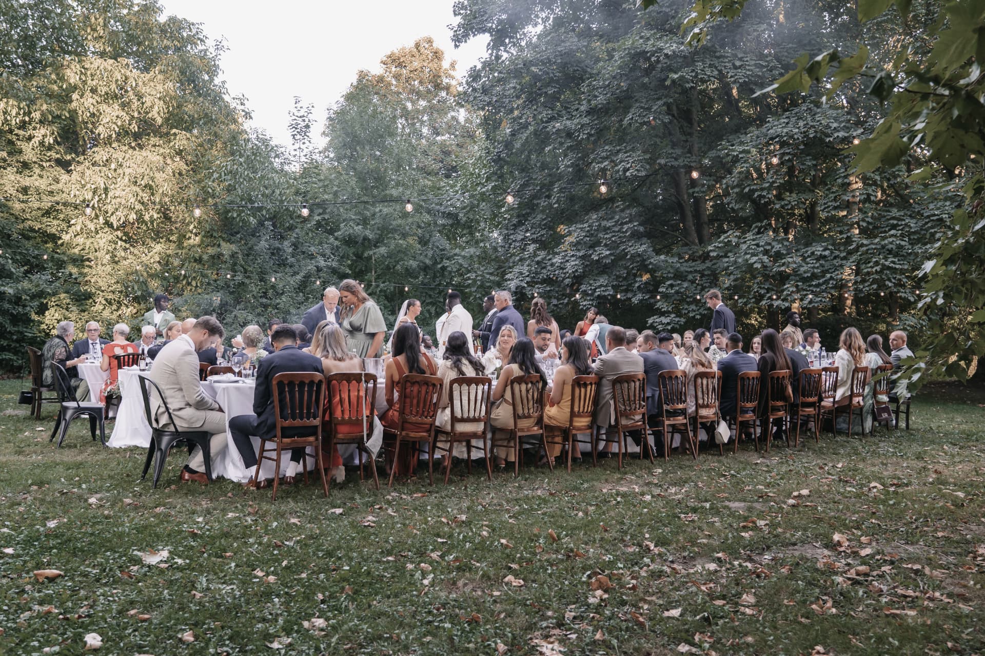 Lange Hochzeitstafel im Grünen mit Gästen beim Dinner unter Lichterketten