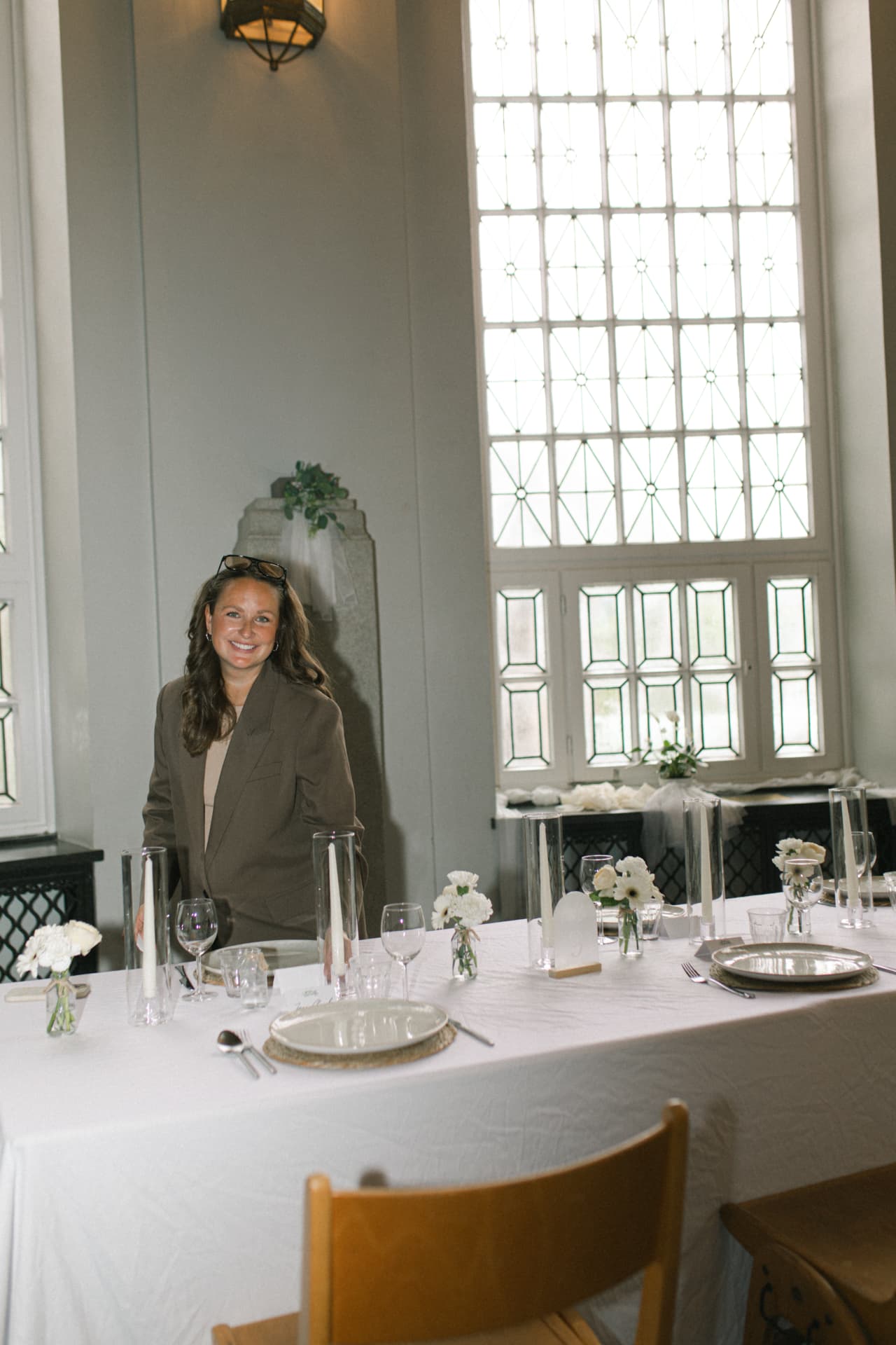 Vivien Opoku steht lächelnd hinter einer festlich gedeckten Hochzeitstafel mit Kerzen, Gläsern und weißen Blumen in einem hellen Raum mit hohen Fenstern
