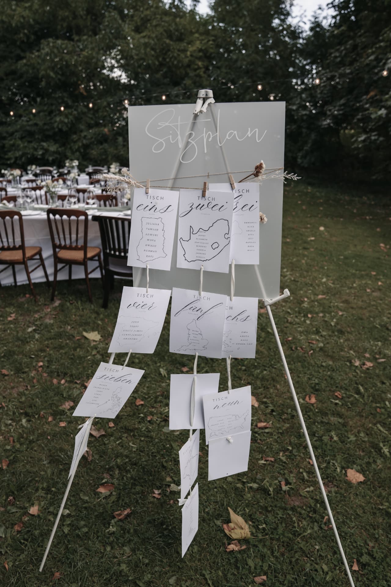 Sitzplan-Tafel auf einer Staffelei mit an Schnüren hängenden Tischkarten vor dem Hochzeitstafel