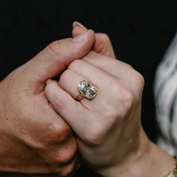Zwei ineinander verschränkte Hände, an einem Finger Taylor Swifts goldener Verlobungsring mit großem Diamanten im Kissenschliff
