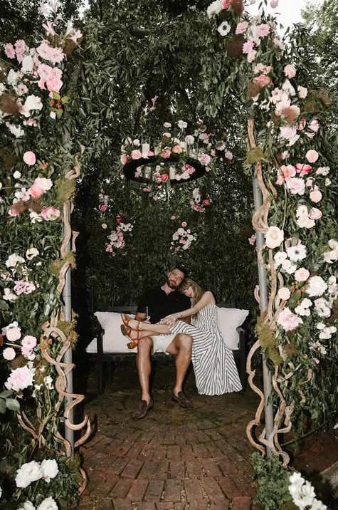 Taylor Swift und Travis Kelce entspannt auf einer Bank unter einem mit rosa und weißen Rosen bewachsenen Laubengang