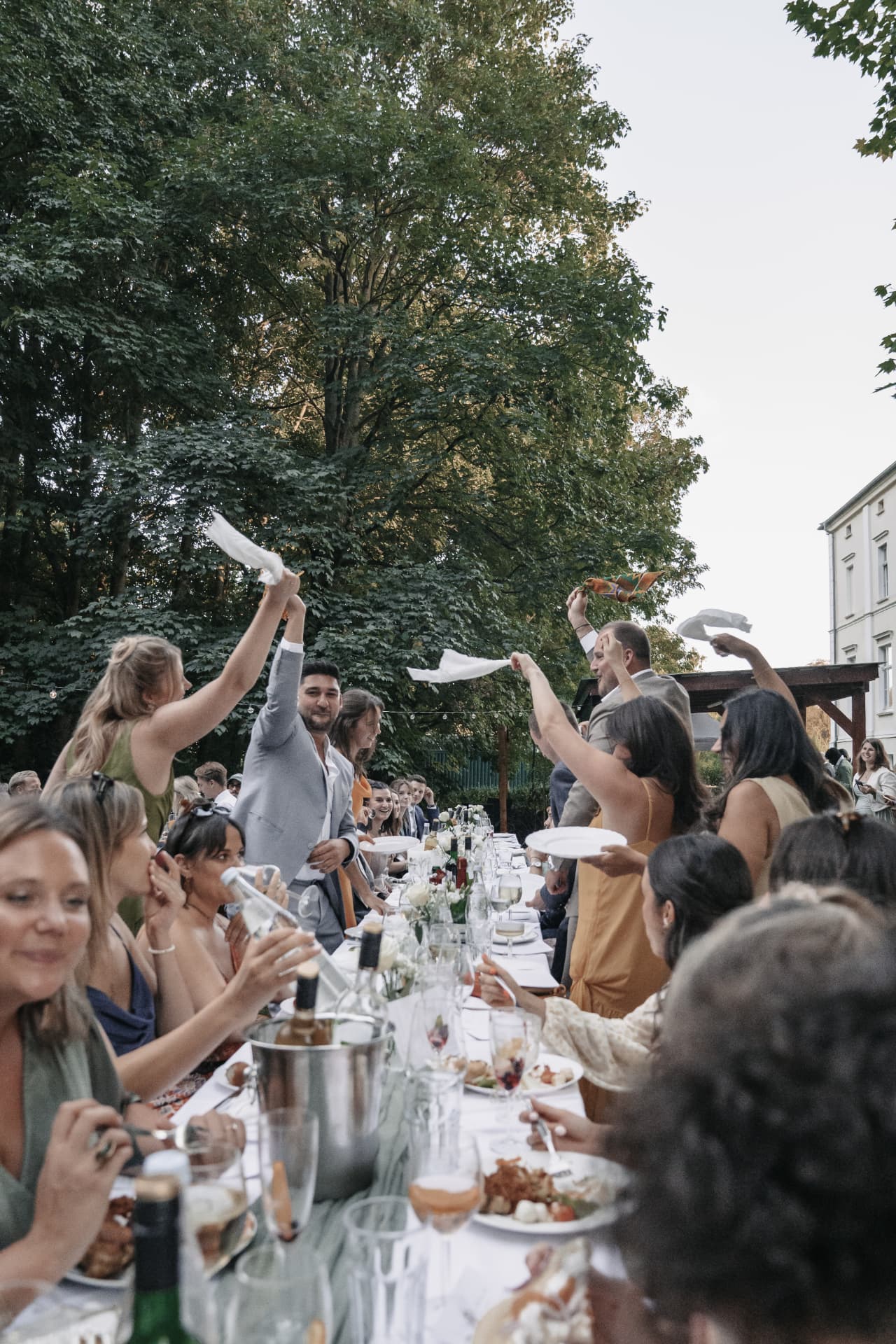 Gäste feiern an einer langen Tafel im Freien und erheben die Servietten